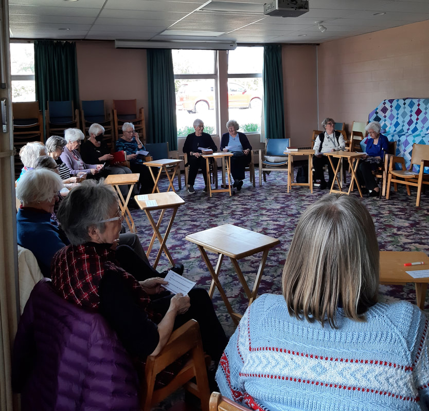 A photograph taken in the Friendship Room of the Caroline Harcourt Women's Friendship Circle, taken in April 2023. The group is sitting in seats arranged in a circle and are enjoying conversation and treats.