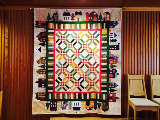 A photograph of a quilt made by the Quilting Club. It hangs in our Sanctuary and depicts a diverse, colourful, and lively neighbourhood where all can find belonging.